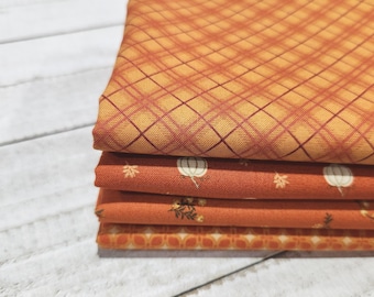 May include: A stack of four folded fabric pieces in shades of orange and gold. The top piece features a diagonal plaid pattern, while the middle layers display small white pumpkin and autumn leaf motifs, and the bottom layer has a geometric pattern.
