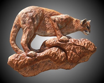 May include: A wooden carving of a cougar standing on a rock. The cougar is a reddish-brown color and has a detailed, realistic appearance. The carving is a work of art by Richard Vest - Rising Tide Sculpture.