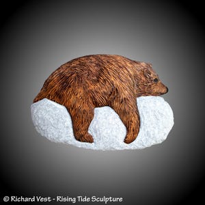 May include: A wooden sculpture of a brown bear resting on a gray rock. The bear is carved in detail, with fur and facial features visible. The sculpture is a realistic depiction of a bear in a relaxed pose.