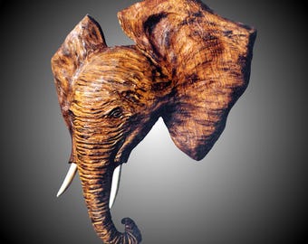 Könnte beinhalten: Eine Holzskulptur eines Elefantenkopfes, detailliert und realistisch geschnitzt. Die Skulptur ist in Brauntönen bemalt und hat eine natürliche Holzmaserung. Der Rüssel des Elefanten ist lang und gebogen, und seine Ohren sind groß und schlaff. © Richard Vest - Rising Tide Sculpture