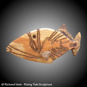 May include: A wooden sculpture of a fish with intricate details carved into the wood. The fish is brown and has a light brown stripe running down its side. The sculpture is made of wood and has a natural finish.