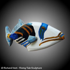 May include: A handcrafted sculpture of a fish with a white body, accented with blue, black, and orange details. The fish has detailed fins and a textured surface. The sculpture is a decorative art piece.