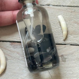 May include: A clear glass spray bottle filled with dark liquid and small black stones. A white tooth is next to the bottle on a light wood surface. The bottle has a black cap and a white spray nozzle.
