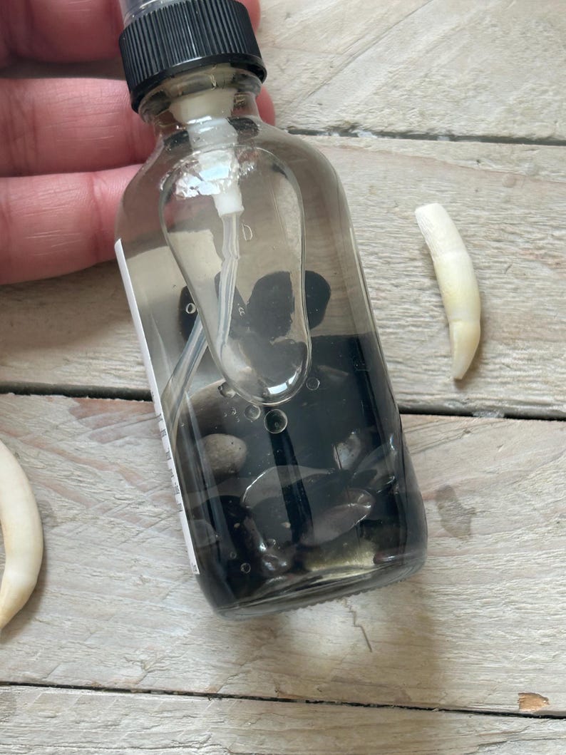 May include: A clear glass spray bottle filled with dark liquid and small black stones. A white tooth is next to the bottle on a light wood surface. The bottle has a black cap and a white spray nozzle.
