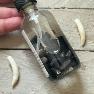 May include: A clear glass spray bottle with a black cap, filled with dark liquid and small black stones. Two off-white, curved objects are next to the bottle. The bottle has a white label with text.
