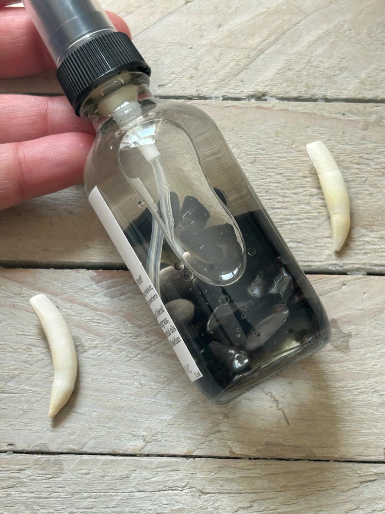 May include: A clear glass spray bottle with a black cap, filled with dark liquid and small black stones. Two off-white, curved objects are next to the bottle. The bottle has a white label with text.
