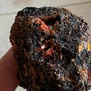 Könnte beinhalten: Ein rauer, dunkelbrauner Stein mit einem Cluster von leuchtend orangefarbenen Kristallen, die darin eingebettet sind. Die Kristalle sind scharf und spitz und bilden einen starken Kontrast zum dunklen Hintergrund.