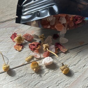 May include: A black pouch spills out bath salts, dried flowers, and rose petals onto a weathered, light-colored wooden surface. The salts are a mix of pink and clear crystals, with small, dried chamomile flowers scattered around.