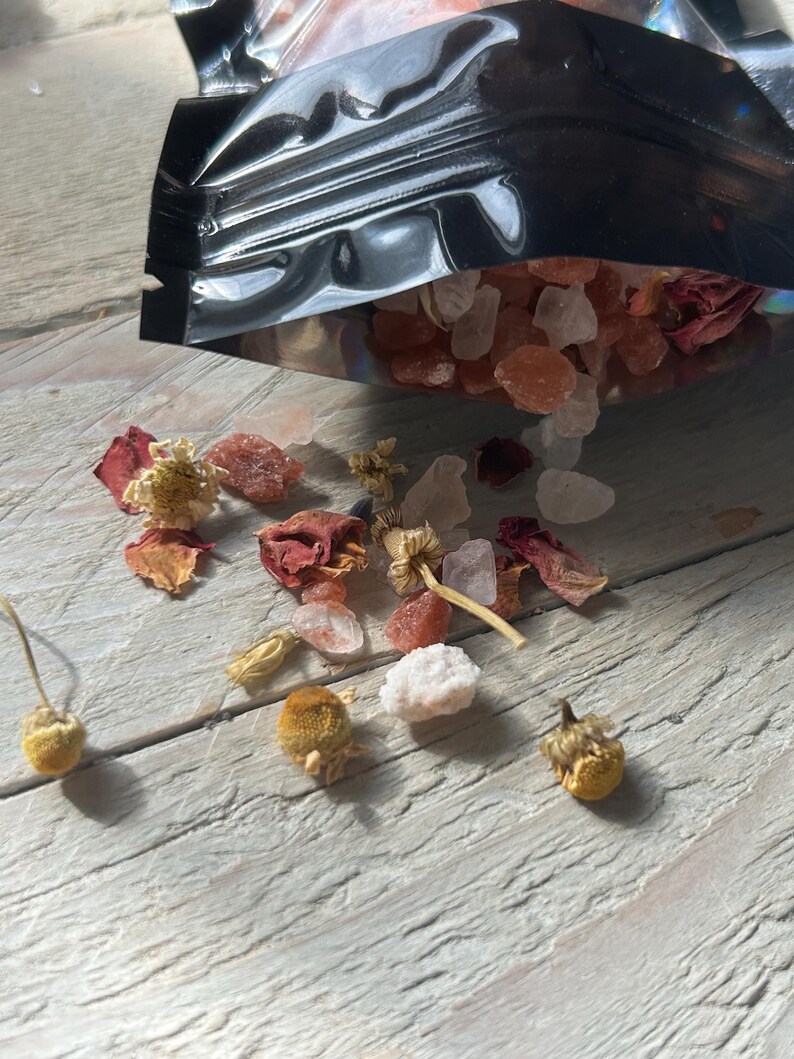 May include: A black pouch spills out bath salts, dried flowers, and rose petals onto a weathered, light-colored wooden surface. The salts are a mix of pink and clear crystals, with small, dried chamomile flowers scattered around.