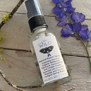 May include: A clear glass bottle with a black spray nozzle and a white label that reads "I AM WILD Natural Room Spray A woodsy blend of Organic Redwood, Patchouli, Sandalwood and Linden Flower essential oils in Forest Rhyolite and Quartz charged Spring Water SACRED WILD DIVINE 1 oz".