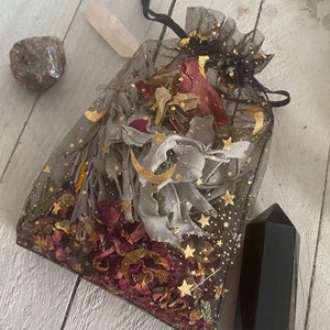 May include: A black mesh bag filled with dried herbs, flowers, and gold glitter stars. The bag is tied with a black ribbon and sits on a white wooden surface. A clear quartz crystal and a black obsidian point are also visible.