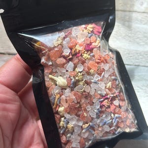 May include: A clear, resealable black pouch filled with bath salts. The salts are a mix of pink, white, and clear crystals, with dried flower petals and lavender buds. The pouch is held in a person's hand.