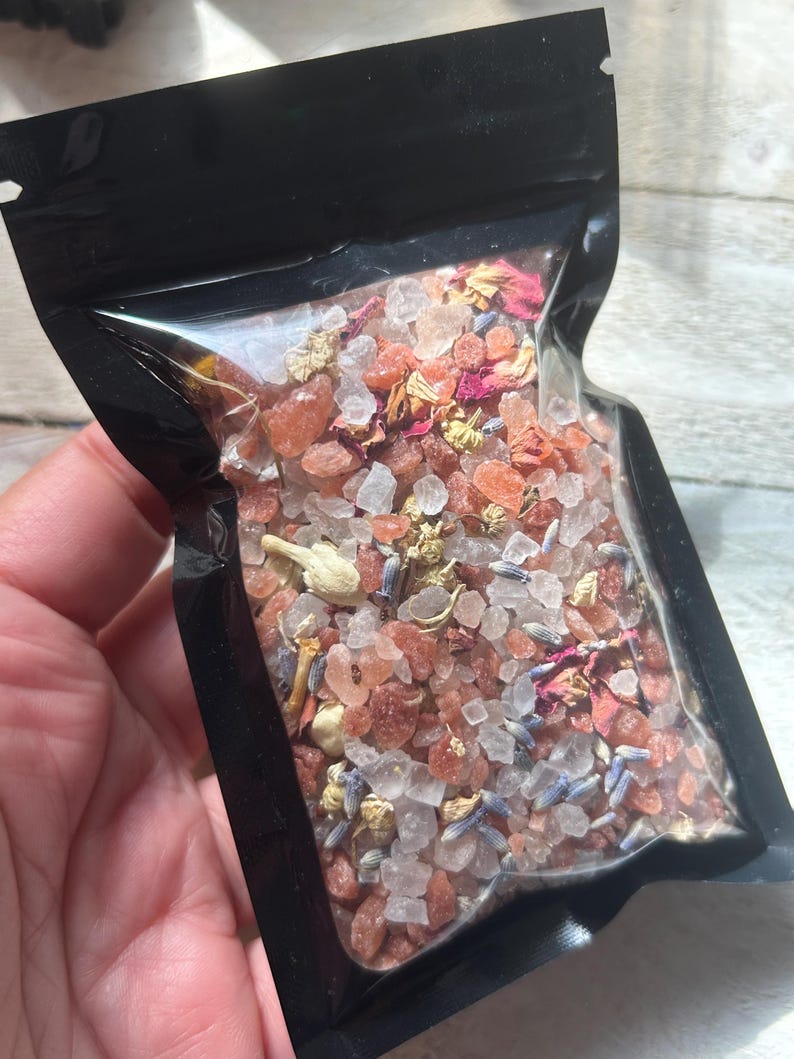 May include: A clear, resealable black pouch filled with bath salts. The salts are a mix of pink, white, and clear crystals, with dried flower petals and lavender buds. The pouch is held in a person's hand.