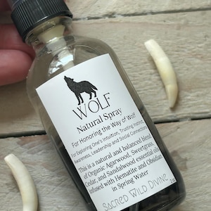 May include: A clear glass spray bottle with a black cap and a white label featuring a wolf silhouette and the words "WOLF Natural Spray." The bottle contains a dark liquid and is accompanied by two white, curved objects. The label includes text about the spray's purpose and ingredients.