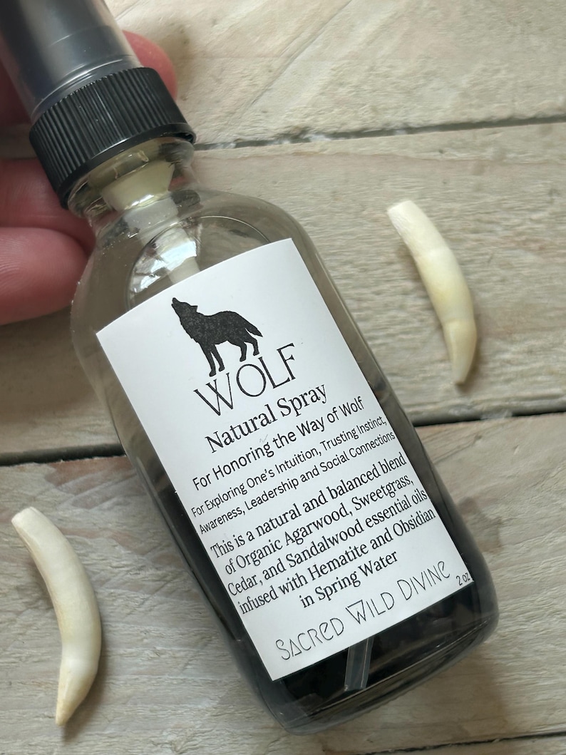May include: A clear glass spray bottle with a black cap and a white label featuring a wolf silhouette and the words "WOLF Natural Spray." The bottle contains a dark liquid and is accompanied by two white, curved objects. The label includes text about the spray's purpose and ingredients.