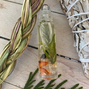 May include: A clear glass roller bottle with a black cap filled with a light yellow liquid and green and orange botanicals. The bottle is sitting on a light wood surface with a braided green and brown cord and a white sage smudge stick.