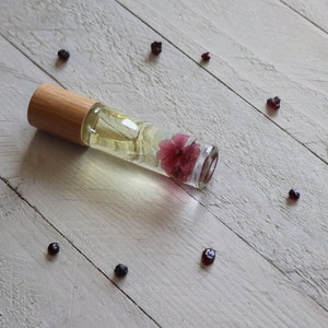 May include: A clear glass roller bottle with a wooden cap and a pink flower inside. The bottle is lying on a white wooden surface with small dark red berries scattered around it.