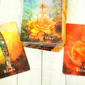 May include: A deck of oracle cards with colorful illustrations. The cards feature images of a lotus flower, a blade, and a heart. The text on the cards includes "Beauty Way", "The Blade", and "The Beloved".