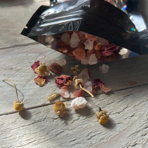 May include: A black pouch partially open, spilling out a mix of pink and white crystals, dried rose petals, and small yellow flowers. The items are scattered on a light-colored wooden surface, creating a natural, spa-like aesthetic.