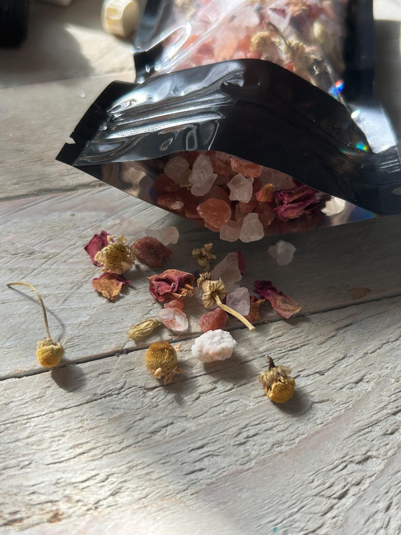 May include: A black pouch partially open, spilling out a mix of pink and white crystals, dried rose petals, and small yellow flowers. The items are scattered on a light-colored wooden surface, creating a natural, spa-like aesthetic.