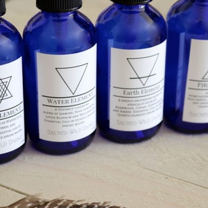 May include: Five blue glass bottles with white labels and geometric symbols. Each bottle is labelled with an element: Aether, Water, Earth, Fire, and Air. The labels also include the text "Sacred Wild Divine".