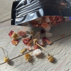 May include: A black metallic bag partially open, spilling out pink and white salt crystals, dried rose petals, and small yellow flowers. The contents are scattered on a light gray wooden surface.