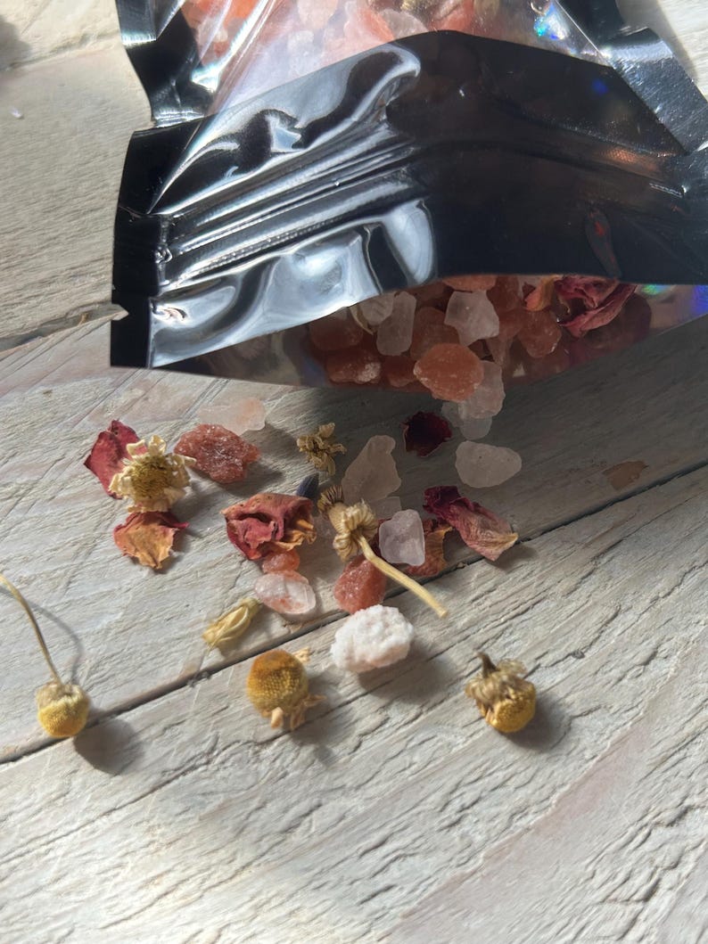 May include: A black metallic bag partially open, spilling out pink and white salt crystals, dried rose petals, and small yellow flowers. The contents are scattered on a light gray wooden surface.