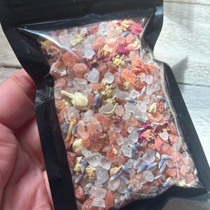 May include: A clear plastic bag filled with bath salts. The salts are a mix of pink and white crystals, with dried lavender, rose petals, and other botanicals. The bag is held in a hand.