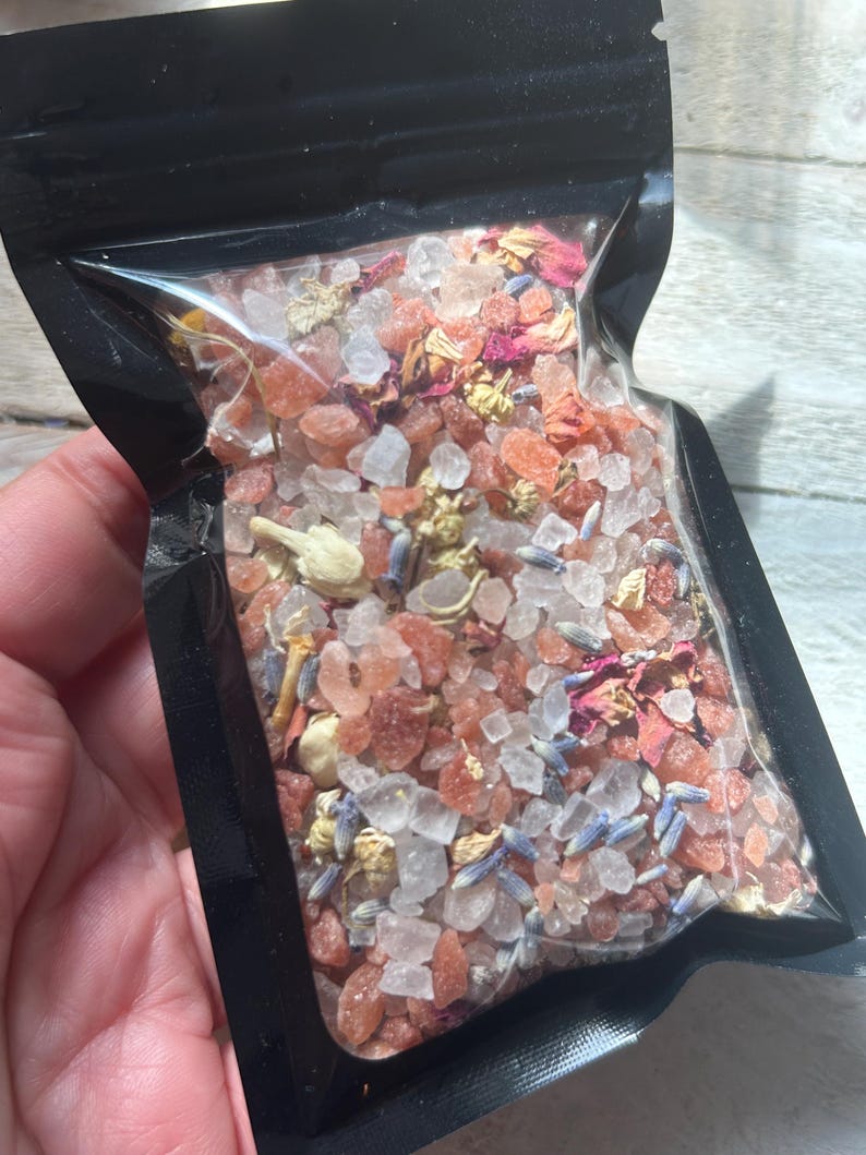 May include: A clear plastic bag filled with bath salts. The salts are a mix of pink and white crystals, with dried lavender, rose petals, and other botanicals. The bag is held in a hand.