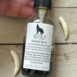 May include: A clear glass spray bottle with a black cap and a white label featuring a black wolf silhouette. The label reads "WOLF Natural Spray" and lists ingredients. Two off-white, curved objects are next to the bottle. The bottle contains a dark liquid.