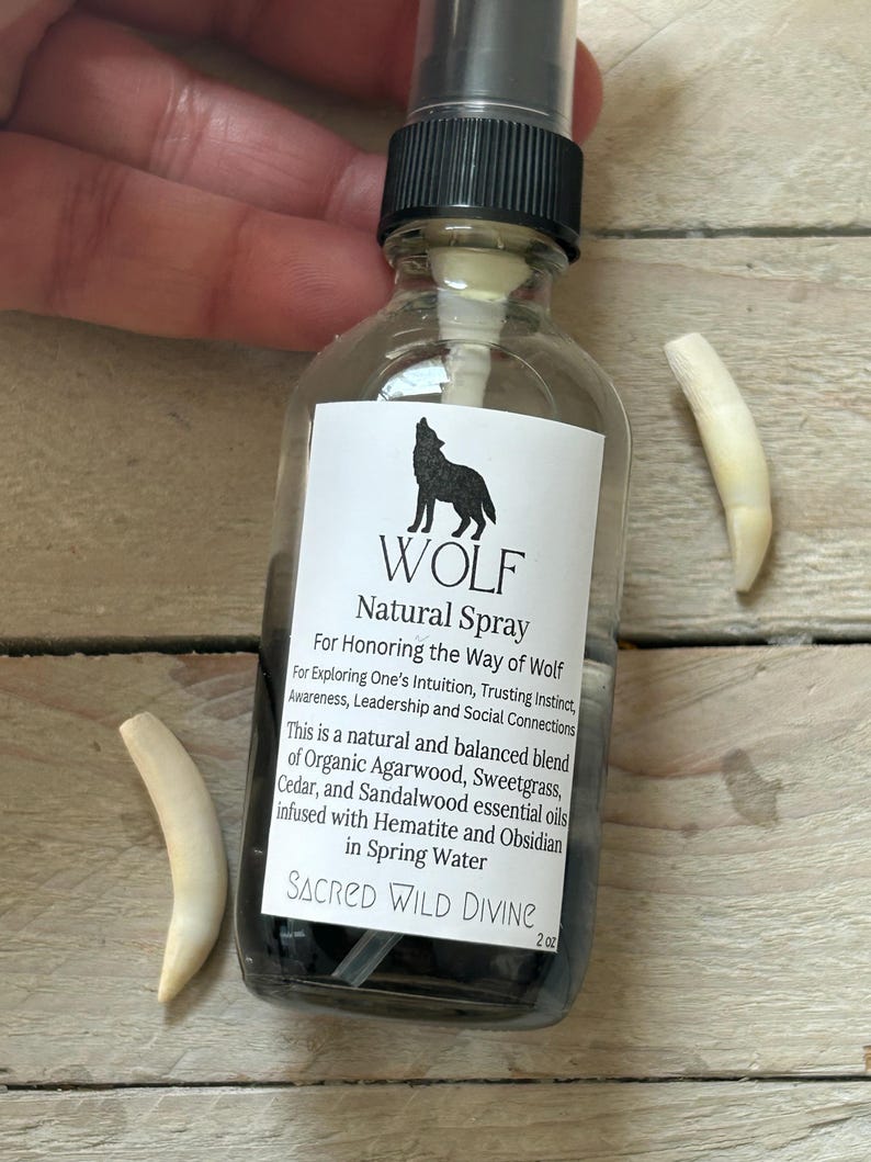 May include: A clear glass spray bottle with a black cap and a white label featuring a black wolf silhouette. The label reads "WOLF Natural Spray" and lists ingredients. Two off-white, curved objects are next to the bottle. The bottle contains a dark liquid.