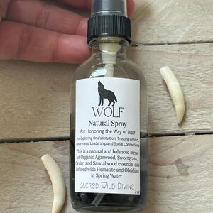 May include: A clear glass spray bottle with a black cap and a label that reads "WOLF Natural Spray." The label features a silhouette of a howling wolf. Two off-white, curved objects are on either side of the bottle. The bottle contains a dark liquid.