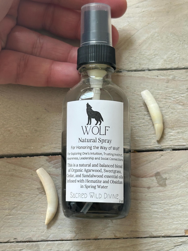 May include: A clear glass spray bottle with a black cap and a label that reads "WOLF Natural Spray." The label features a silhouette of a howling wolf. Two off-white, curved objects are on either side of the bottle. The bottle contains a dark liquid.
