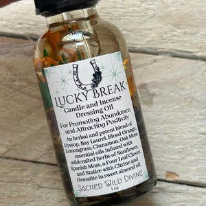 May include: A clear glass bottle with a black cap, labeled "LUCKY BREAK Candle and Incense Dressing Oil." The label features a horseshoe and floral design. The bottle contains herbs and oils, with text promoting abundance and positivity. The bottle is 1 oz.