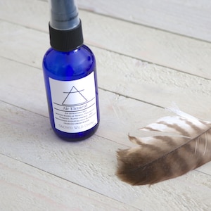 May include: A blue glass bottle with a black spray top and a white label that reads "Air Element". The bottle is sitting on a white wooden surface. A brown and white feather is lying on the surface to the right of the bottle.
