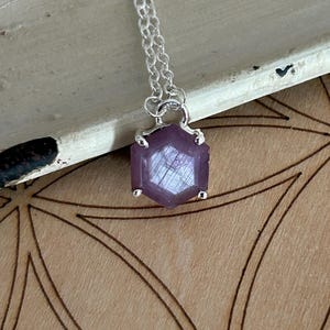 May include: A silver necklace featuring a hexagonal, faceted, purple gemstone pendant. The pendant is set in a silver bezel and hangs from a delicate silver chain. The gemstone has a translucent quality, and the necklace is displayed on a wooden surface.