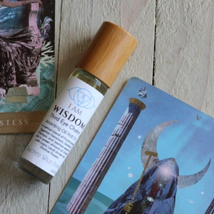 以下が含まれることがあります： 木製のキャップが付いたガラス瓶に、青と白のラベルが貼られています。ラベルには「I AM WISDOM Third Eye Chakra Anointing Oil Roll On SACRED WILD SOUL」と書いてあります。瓶は木製の表面に置かれており、その隣には2枚のタロットカードがあります。1枚は「HIGH PRIESTESS」と書かれており、もう1枚は三日月と2本の柱が描かれています。