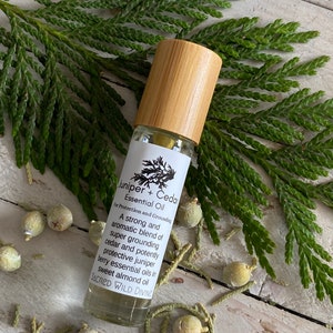 May include: A glass roller bottle with a wooden cap, filled with a clear liquid. The label reads "Juniper + Cedar Essential Oil for Protection and Grounding. A strong and aromatic blend of super grounding cedar and potently protective juniper berry essential oils in sweet almond oil. SACRED WILD DIVINE."