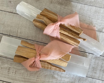 Può includere: Due mazzi di bastoncini di Palo Santo avvolti in tessuto rosa, ogni mazzo è posto sopra un cristallo di selenite bianco.