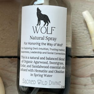 May include: A clear glass bottle with a black liquid and a white label. The label features a black silhouette of a howling wolf above the word "WOLF" and the text "Natural Spray". The bottle contains 2 oz of product.