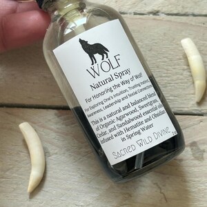 May include: A clear glass spray bottle with a black cap and a white label featuring a black wolf silhouette and the word "WOLF". The label also includes text about the product's purpose and ingredients. Two white, curved objects are next to the bottle.