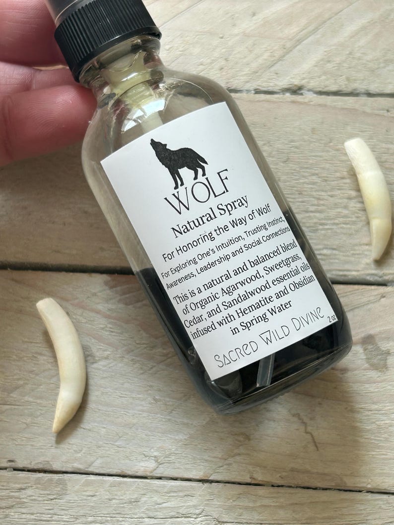 May include: A clear glass spray bottle with a black cap and a white label featuring a black wolf silhouette and the word "WOLF". The label also includes text about the product's purpose and ingredients. Two white, curved objects are next to the bottle.