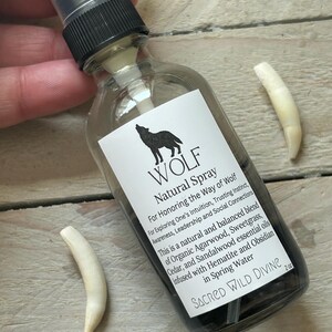 May include: A clear glass spray bottle with a black cap and a white label featuring a wolf silhouette and text. The label reads "WOLF Natural Spray." Two off-white, curved objects are next to the bottle. The bottle contains a dark liquid.