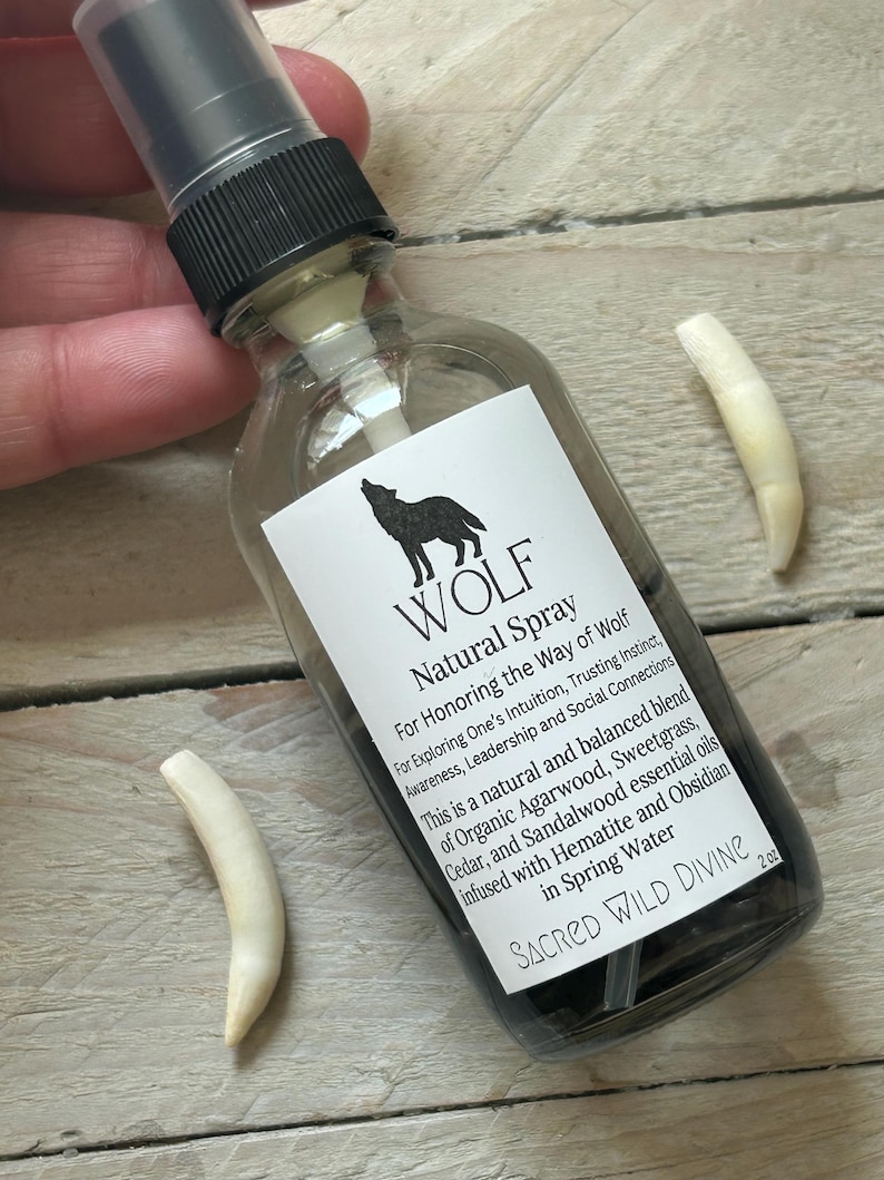 May include: A clear glass spray bottle with a black cap and a white label featuring a wolf silhouette and text. The label reads "WOLF Natural Spray." Two off-white, curved objects are next to the bottle. The bottle contains a dark liquid.