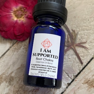 May include: A blue glass bottle with a black cap, labeled "I AM SUPPORTED Root Chakra Essential Oil Blend". The label also includes the text "A supportive blend of Damascus Rose, Sandalwood, Myrrh, and Geranium essential oils in sweet almond oil" and "SACRED WILD DIVINE".