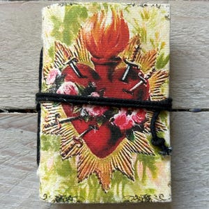 May include: A small, rectangular journal with a heart design. The cover features a red heart pierced with nails, surrounded by roses and a golden sunburst. A black cord wraps around the journal to keep it closed. The background is a mix of green and yellow.
