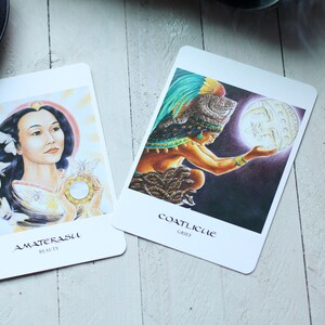 Può includere: Due carte dei tarocchi con illustrazioni e testo. Una carta presenta una donna con lunghi capelli neri e la parola "AMATERASU BEAUTY". L'altra carta mostra una figura che tiene un globo luminoso, con la parola "COATLICUE GRIEF".