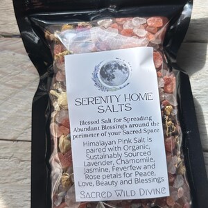 May include: A black pouch filled with pink Himalayan salt and dried flowers. The label reads "SERENITY HOME SALTS" and describes the salt as paired with lavender, chamomile, jasmine, feverfew, and rose petals. The text also mentions "SACRED WILD DIVINE."
