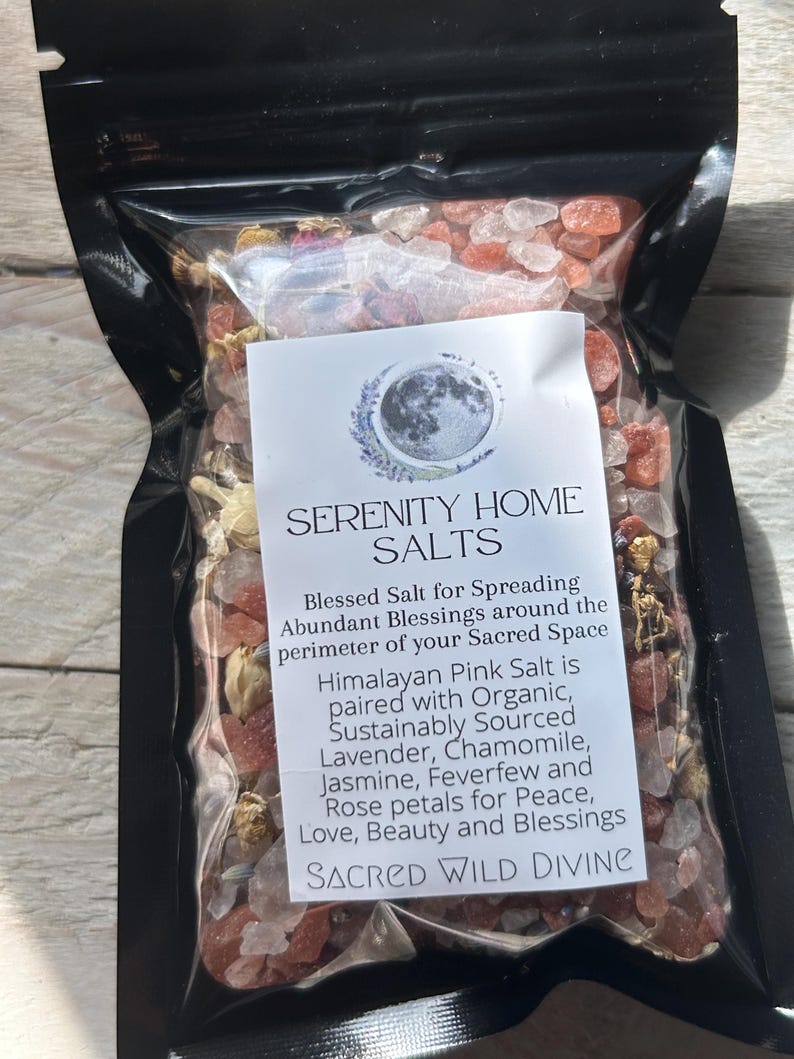 May include: A black pouch filled with pink Himalayan salt and dried flowers. The label reads "SERENITY HOME SALTS" and describes the salt as paired with lavender, chamomile, jasmine, feverfew, and rose petals. The text also mentions "SACRED WILD DIVINE."