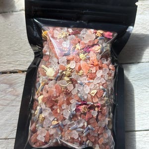 May include: A black, resealable bag filled with a mixture of bath salts. The salts are a mix of pink, white, and clear crystals, with dried lavender and rose petals. The bag is sitting on a white wooden surface.